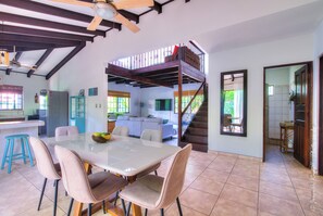 Une salle à manger spacieuse avec un haut plafond et des poutres apparentes, un escalier menant à un niveau supérieur, et une vue sur la cuisine et le salon.