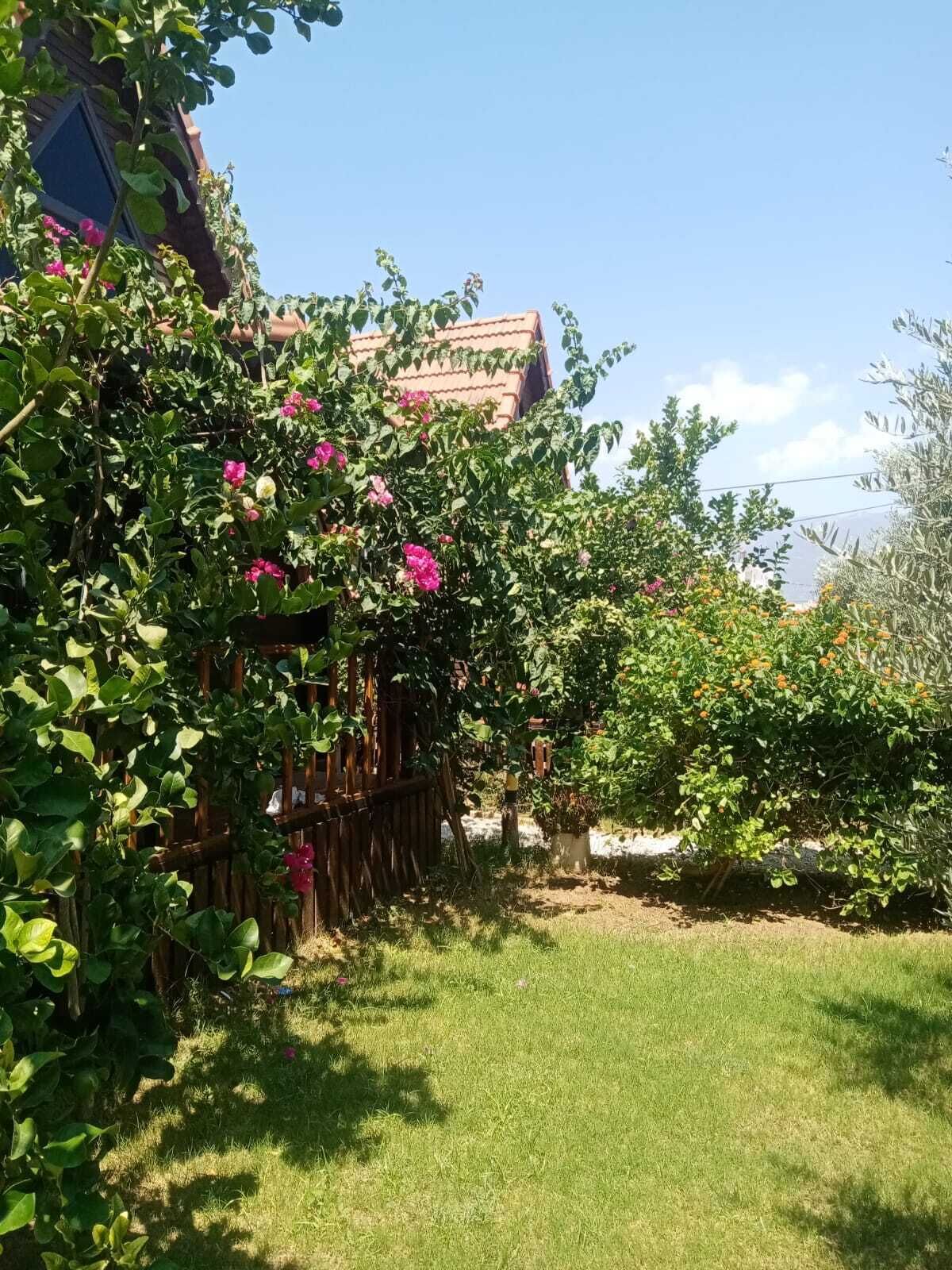 Un jardín con valla de madera, diversas plantas y un edificio con techo de tejas al fondo.