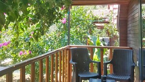 A wooden deck with two wicker chairs, a wooden railing, and a garden with green foliage and pink flowers. - Riviera bungalov apart (Demre)