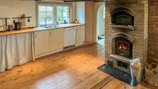 A kitchen with wooden flooring, a brick fireplace, a dishwasher, and a window with a view of greenery.