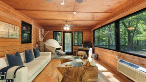 A cabin interior with a wooden ceiling, a ceiling fan, a sofa with pillows, a coffee table, and a large window with a view of trees.