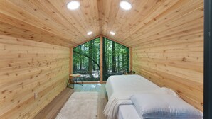 A wooden cabin bedroom with a bed, a chair, and a large window offering a forest view.