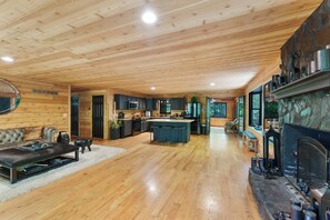Un vaste espace de vie doté d’un plafond en bois, d’une cheminée et d’une cuisine située à l’arrière-plan.