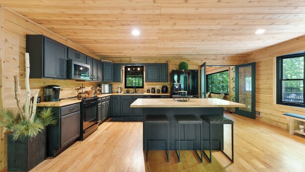 A modern kitchen with wooden flooring, dark cabinetry, a central island with bar stools, and a large window offering a view of the outdoors.