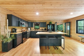A modern kitchen with wooden flooring, dark cabinetry, a central island with bar stools, and a large window offering a view of the outdoors. - Spacious 7-bedroom cabin with AC in charming Stockbridge (Stockbridge)