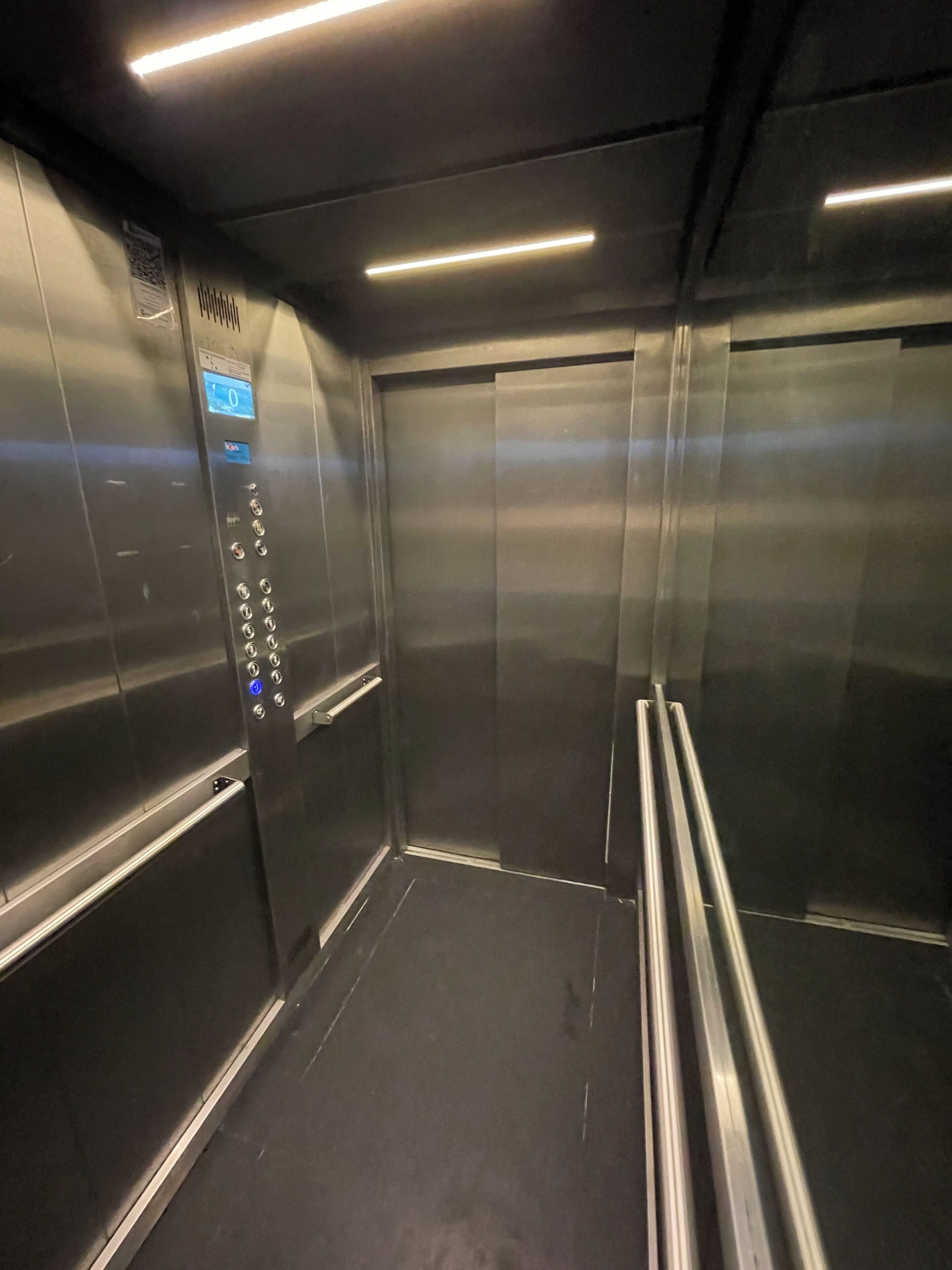 Interior de elevador com paredes metálicas, painel de controle com botões e iluminação no teto.