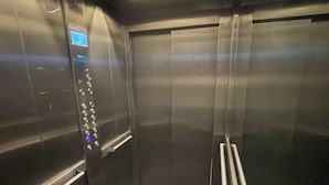 Interno di un ascensore con pareti metalliche, un pannello di controllo con pulsanti e luci a soffitto.