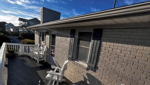 Una terraza techada al aire libre con sillas Adirondack blancas y una pequeña mesa de madera, contigua a un edificio de ladrillo con una ventana y una puerta.