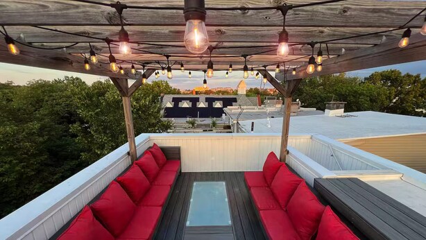Un área de estar al aire libre con cojines rojos y una mesa de vidrio, bajo una pérgola de madera con luces colgantes.