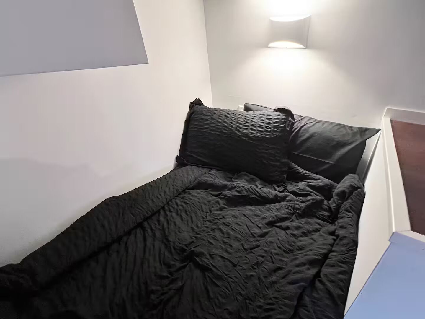 Ein kleines Zimmer mit einem Einzelbett, einer dunkel gefärbten Bettdecke, einem Kopfteil und einer Wandleuchte.