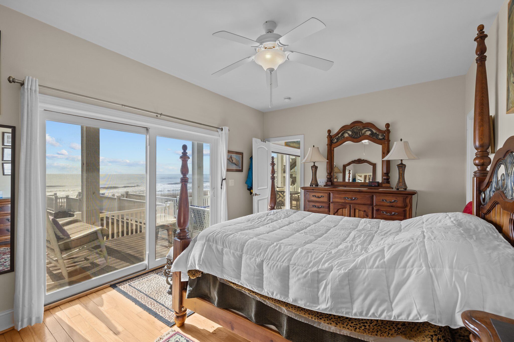 A bedroom with a large bed, a ceiling fan, a wooden dresser, and a view of the beach through a sliding glass door.
