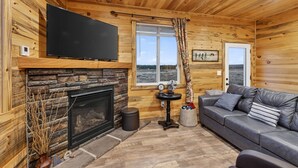 Een gezellig cabin interieur met een stenen open haard, een flatscreen televisie erboven, een grijze bank en een raam met uitzicht op de omgeving.