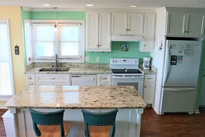 A kitchen with white cabinets, a granite countertop, a refrigerator, and two teal chairs. - 4BR Coastal Retreat with Decks & Grill (Emerald Isle)