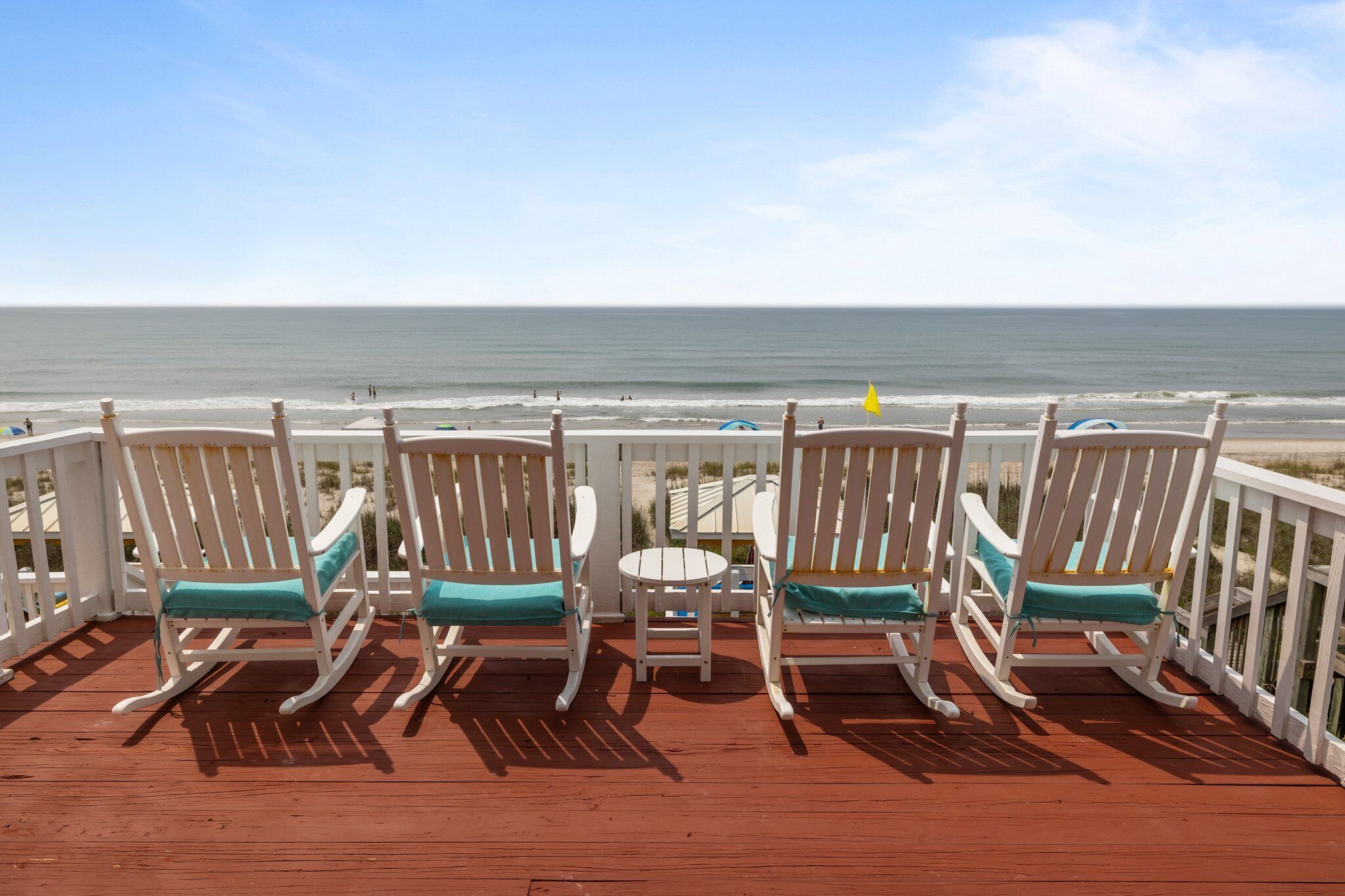 A deck with white rocking chairs and a small table, overlooking a beach.