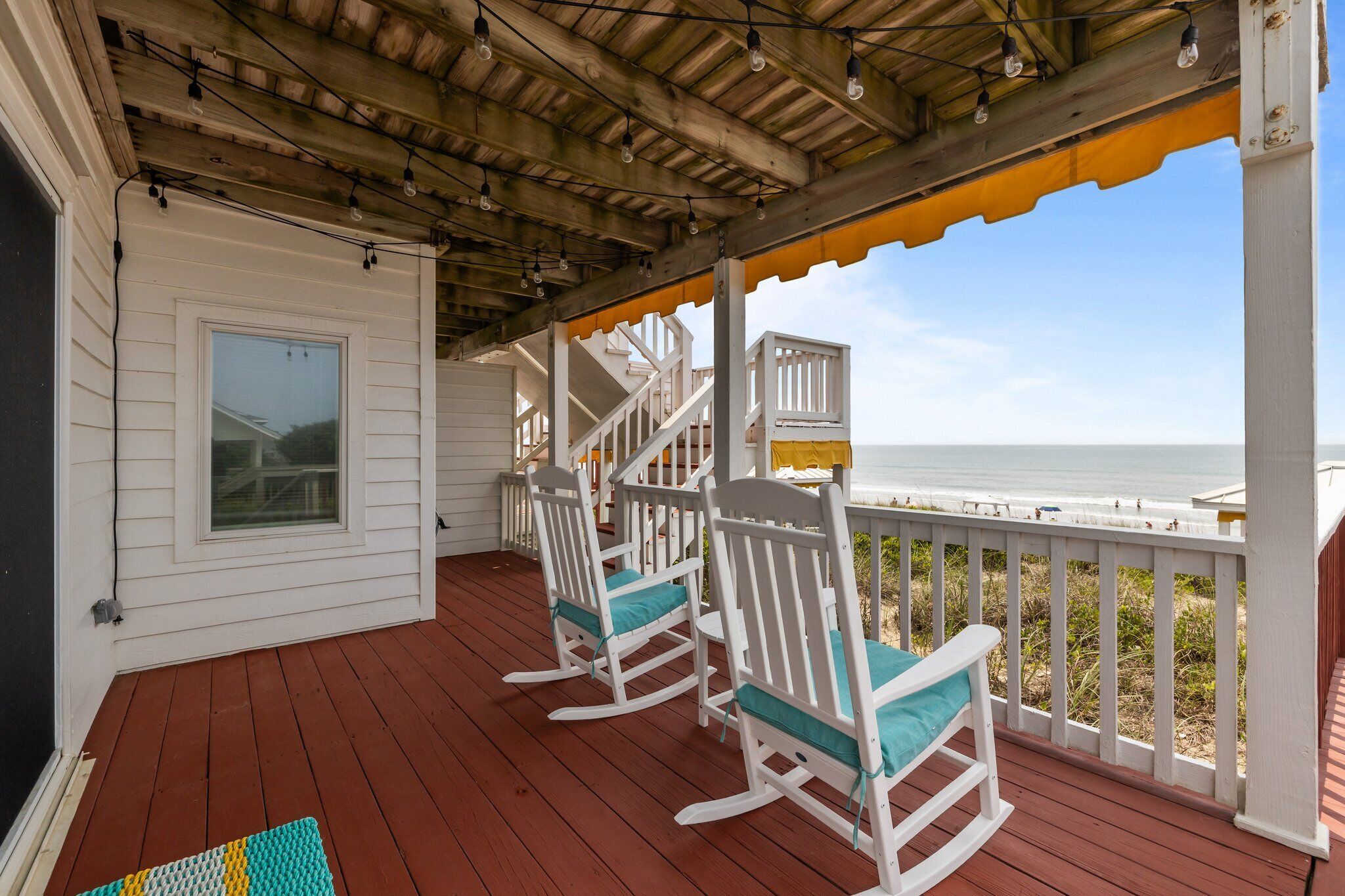 A wooden deck with rocking chairs, a covered area with string lights, and a view of the beach.