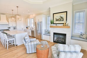 A modern kitchen with white cabinetry, stainless steel appliances, and a central island. There is a dining area with white chairs and a striped sofa. A fireplace with a mantel and framed artwork is visible. - Second Row Ocean View Retreat w/ Pool and Elevator (Emerald Isle)