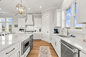 A modern kitchen with white cabinetry, a large island, and a view of the outdoors. - Second Row Ocean View Retreat w/ Pool and Elevator (Emerald Isle)