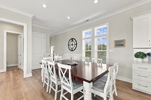 Un coin repas avec une table en bois et des chaises blanches, une horloge au mur et une fenêtre avec des stores.
