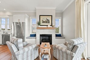 A modern living room with a fireplace, striped sofas, a coffee table, and a kitchen area with a stainless steel refrigerator. - Second Row Ocean View Retreat w/ Pool and Elevator (Emerald Isle)
