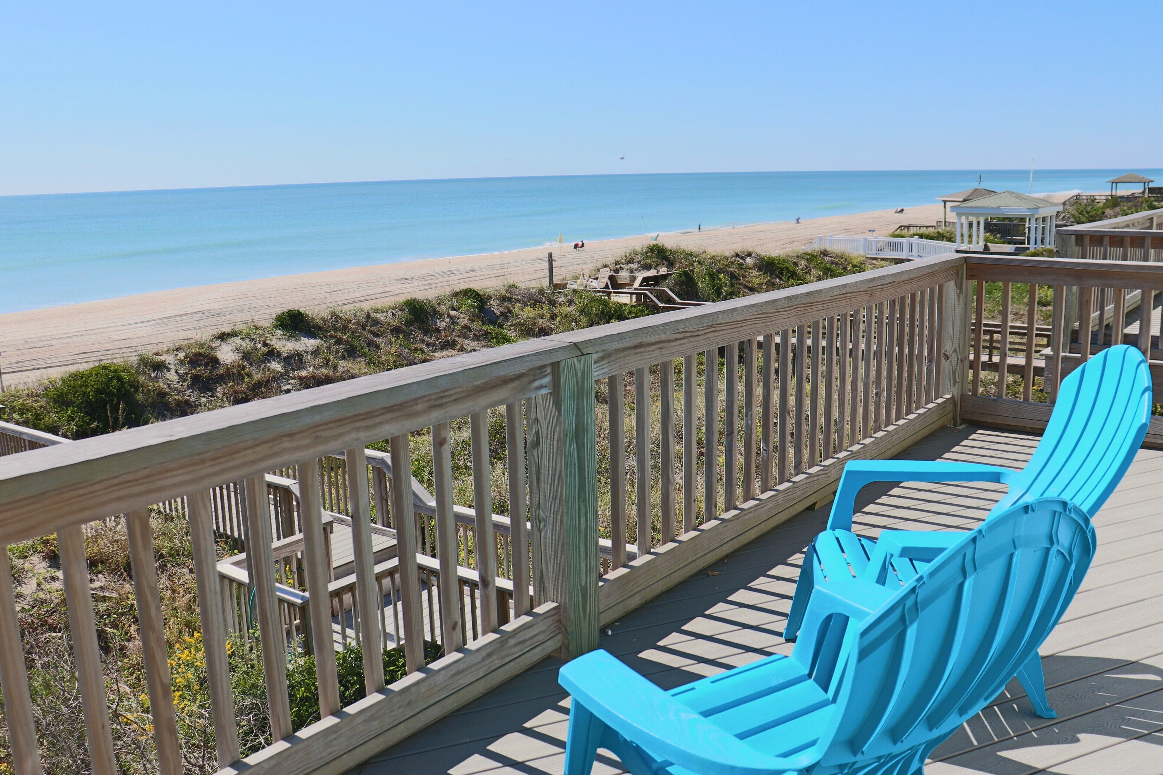 Una terraza de madera con barandilla, dos sillas Adirondack azules y vista a la playa.