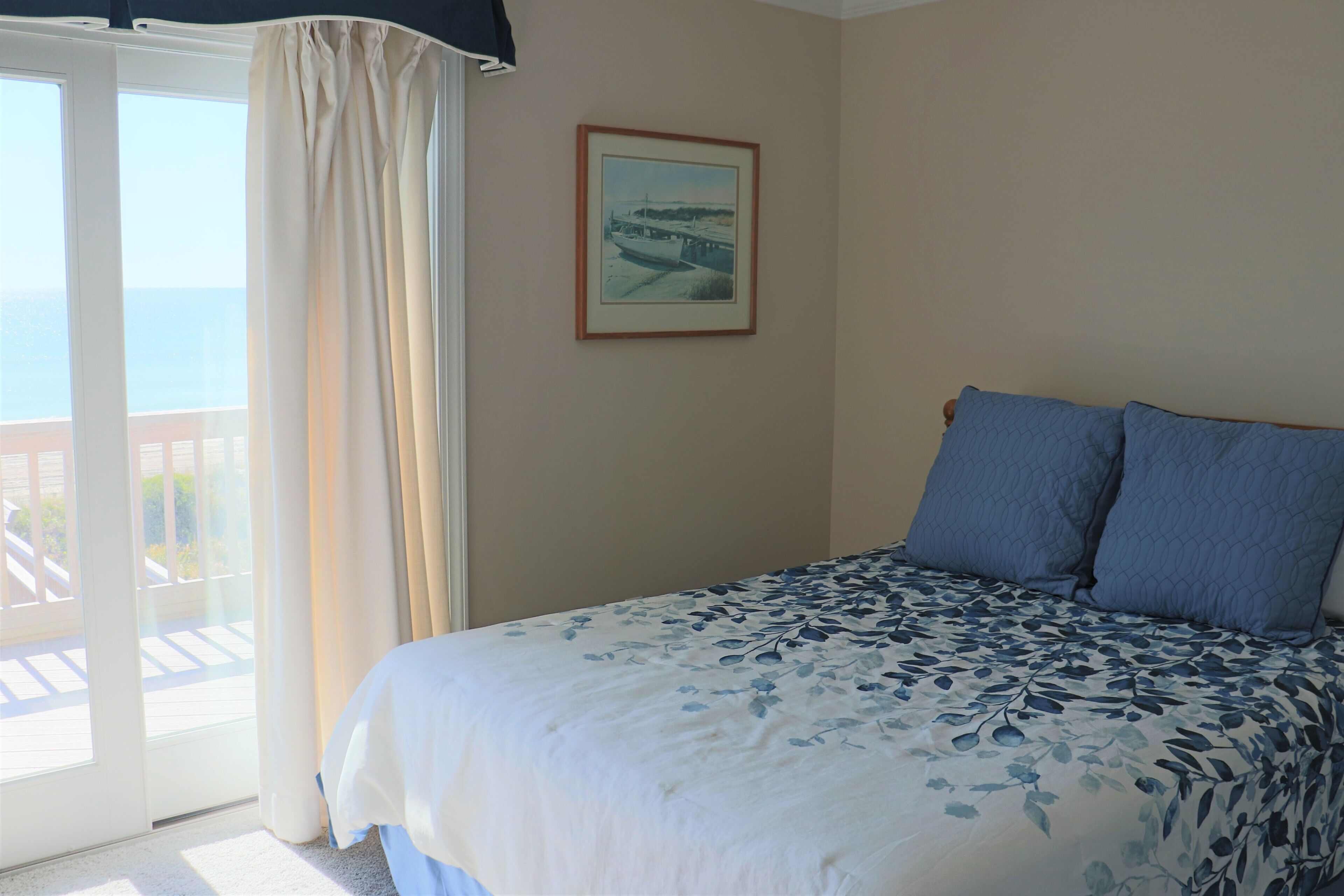 A bedroom with a bed, a window with curtains, a framed picture on the wall, and a view of the outdoors.