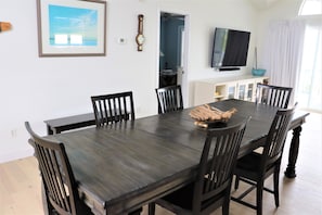 A dining area with a dark wooden table and chairs, a framed picture, a clock, a television, and a cabinet with items on it. - Spinnaker’s Reach Coastal Retreat | Ocean View | Community Pool | Reverse Floor (Emerald Isle)