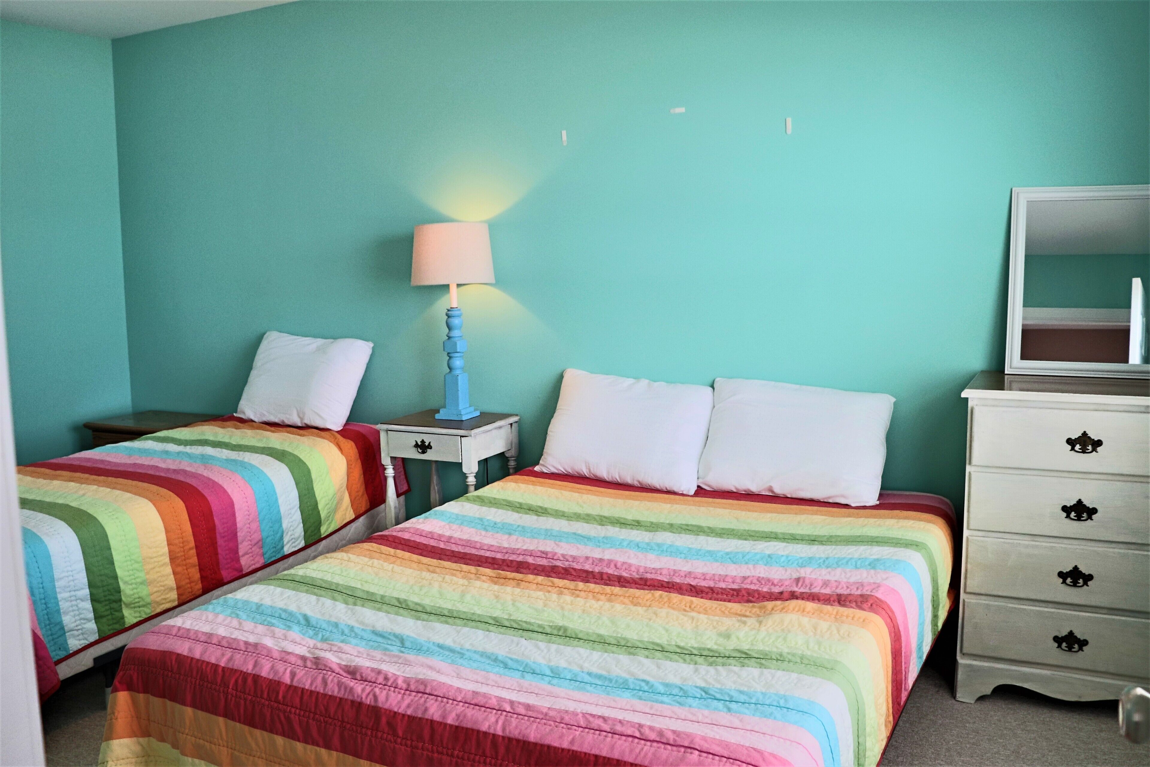 A room with two beds, a nightstand with a lamp, a dresser with drawers, and a mirror.