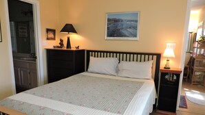 A bedroom with a bed, a nightstand, a lamp, a framed picture, and a door leading to a bathroom. - 3rd Row Home, Elevator & Ocean View (Emerald Isle)
