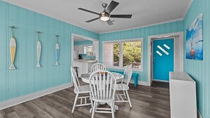 Un coin repas avec des chaises blanches, un ventilateur de plafond et une porte bleue.