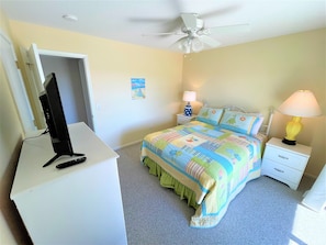 Ein Schlafzimmer mit einem Bett, einem Schreibtisch mit Computer, einem Nachttisch mit Lampe und einem Deckenventilator.