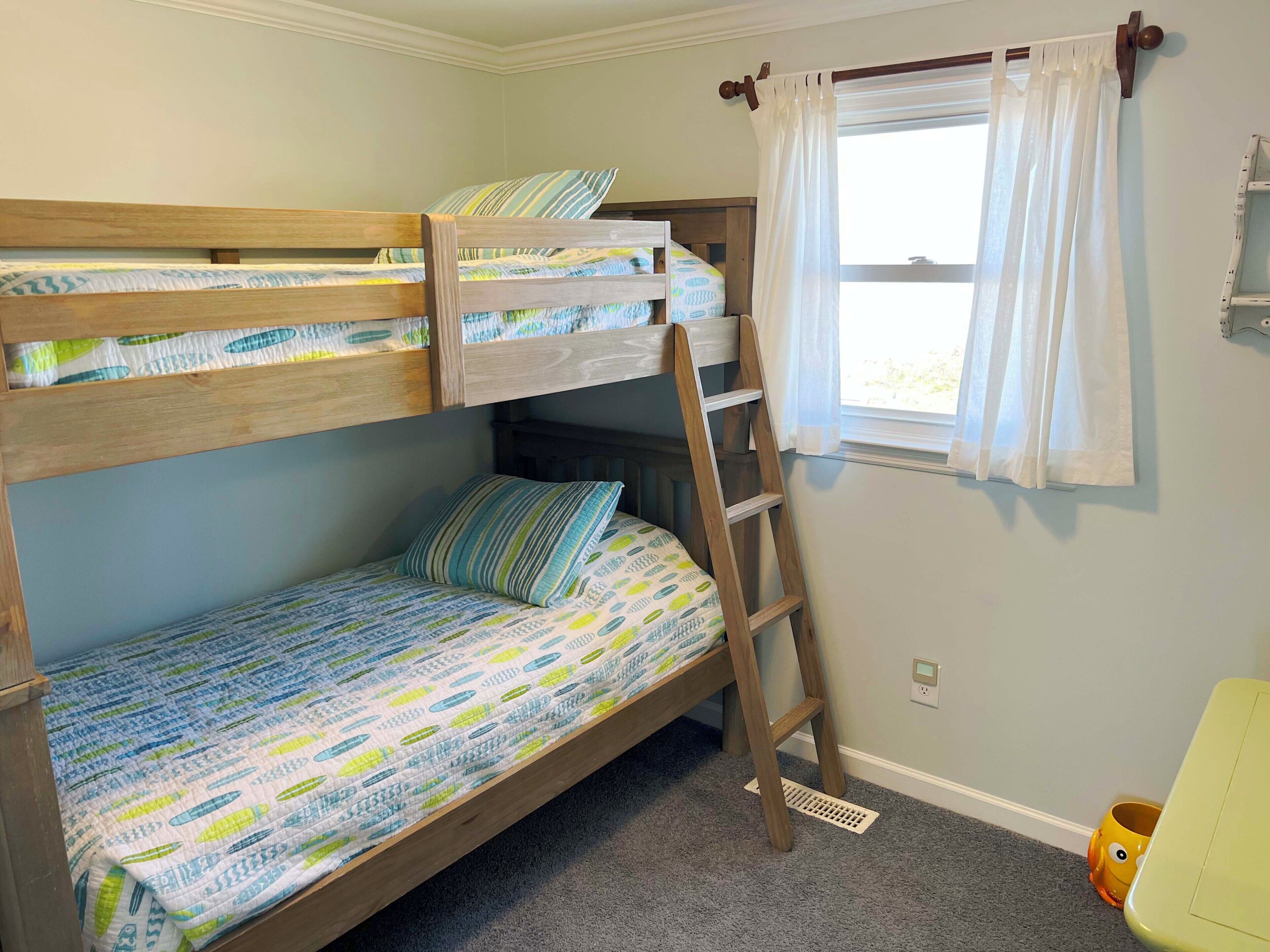 A bunk bed with a ladder, a window with curtains, and a small yellow chair.