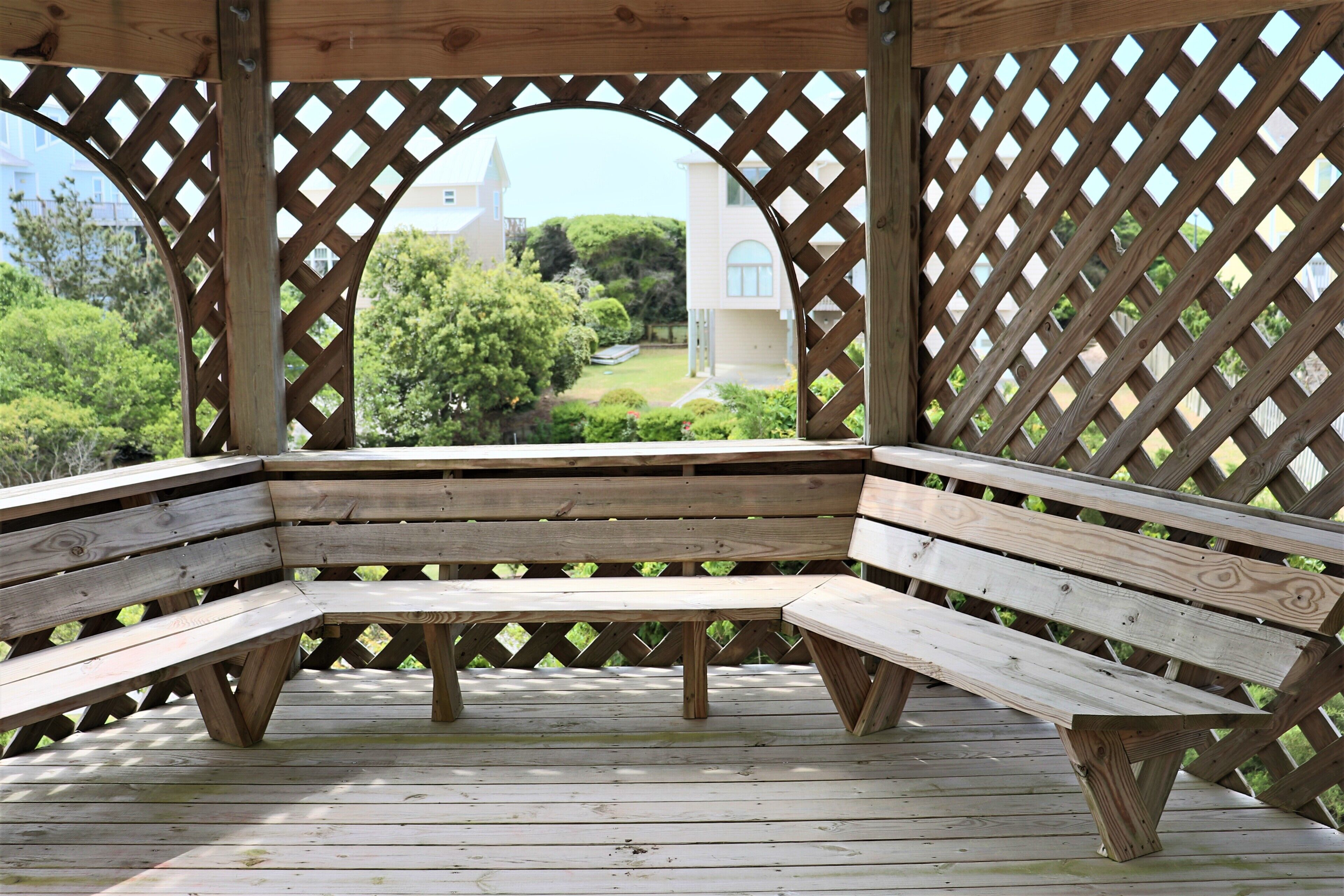 A wooden bench under a wooden lattice structure with a view of a building and greenery.