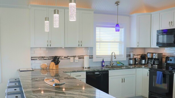 Una cocina moderna con armarios blancos, encimeras de granito, electrodomésticos de acero inoxidable e iluminación colgante.