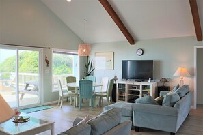 A living room with a TV, sofa, dining table, chairs, and a balcony with outdoor furniture. - Oceanfront 4BR-Decks, Dog-Friendly & Private Beach Walkway (Emerald Isle)