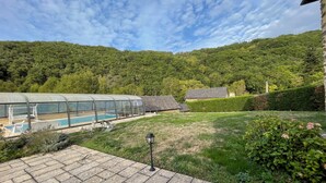 A pool area with a glass enclosure, a stone building, and a lush green hillside in the background. - Cosy country side property with three cottages and covered swimming pool (Centres)