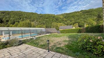 Un espace piscine protégé par une paroi de verre, un bâtiment en pierre et, en arrière-plan, une colline verdoyante.