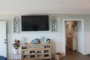 Ein Wohnzimmer mit an der Wand befestigtem Fernseher, einem Holzschrank mit der Aufschrift 'Welcome to the Beach' und einer Zimmerpflanze.