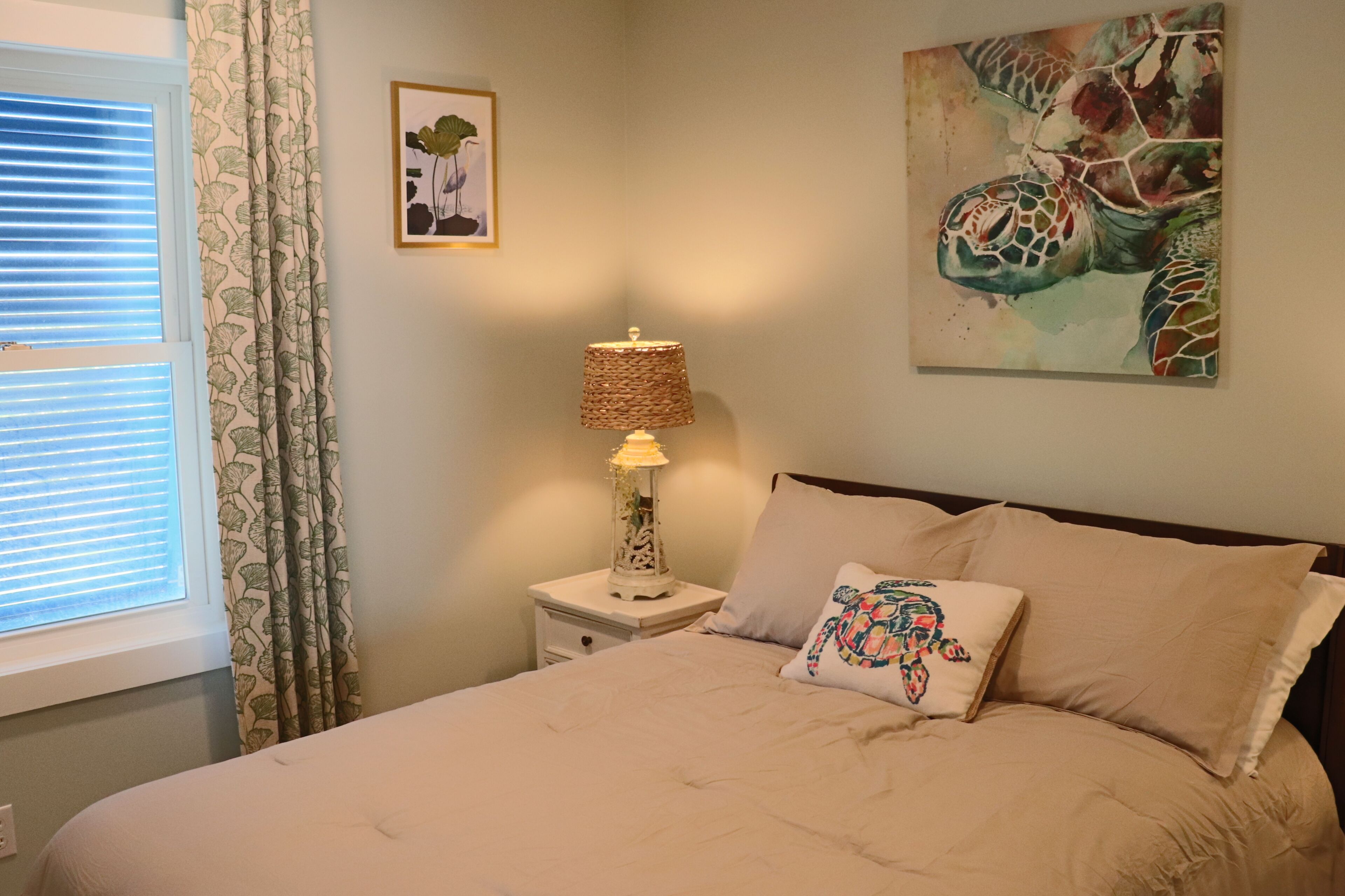 A bedroom with a bed, a nightstand with a lamp, a window with blinds, and a wall with a framed picture.