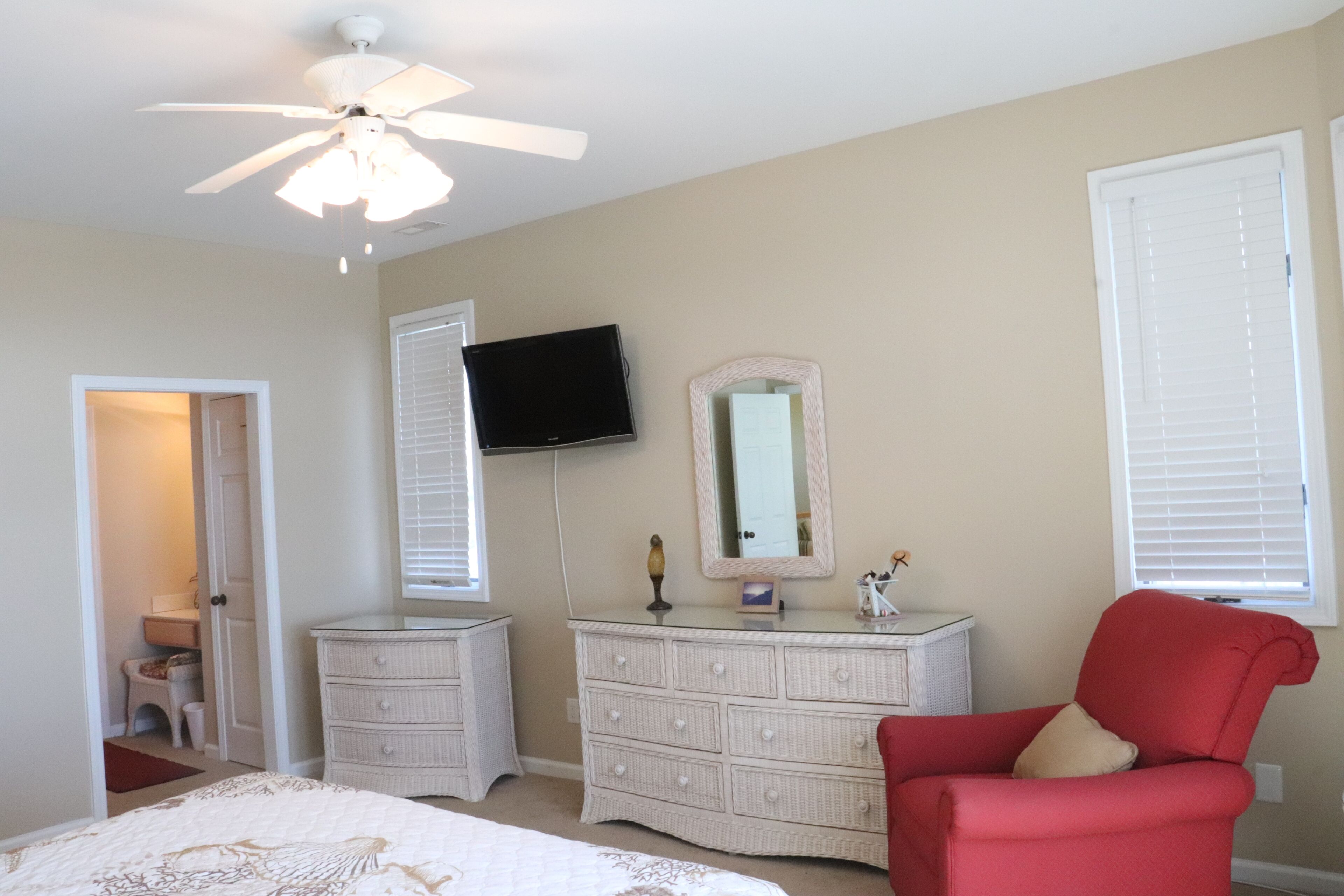 A bedroom with a bed, dresser, chair, ceiling fan, TV, and a door leading to another room.