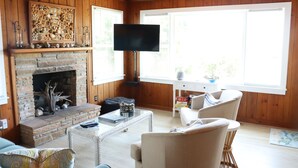 Una sala de estar con chimenea de piedra, televisor de pantalla plana, paredes de madera y ventanas grandes.