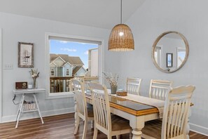 A dining area with a wooden table, white chairs, a wicker pendant light, and a round mirror on the wall. - Family-Friendly Coastal Home | Community Pool | Dog Friendly (Emerald Isle)