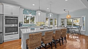 A modern kitchen with stainless steel appliances, a large island with bar stools, and large windows offering views of the outdoors. - Family Retreat with Hot Tub & Game Room and Short Walk to Beach (Emerald Isle)