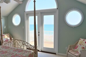 Une chambre à coucher avec un lit à baldaquin, un fauteuil en osier et une vue sur la plage, visible à travers une grande porte.
