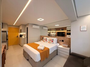 A modern hotel room with a large bed, bedside tables, a built-in TV, a mirror, and a kitchenette. - Condomínio Helbor Edition  (São Paulo)