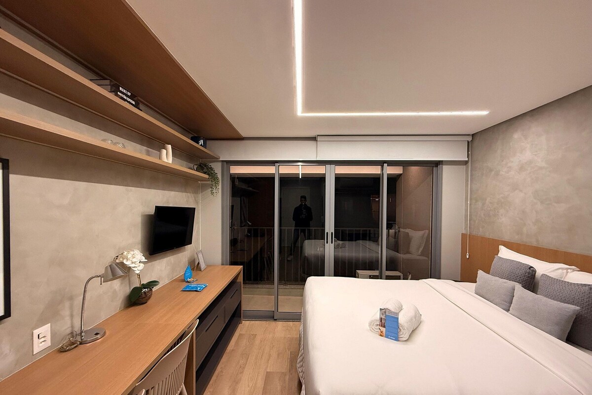 A modern hotel room with a wooden desk, a flat-screen TV, a bed with pillows, and a sliding glass door leading to a balcony.