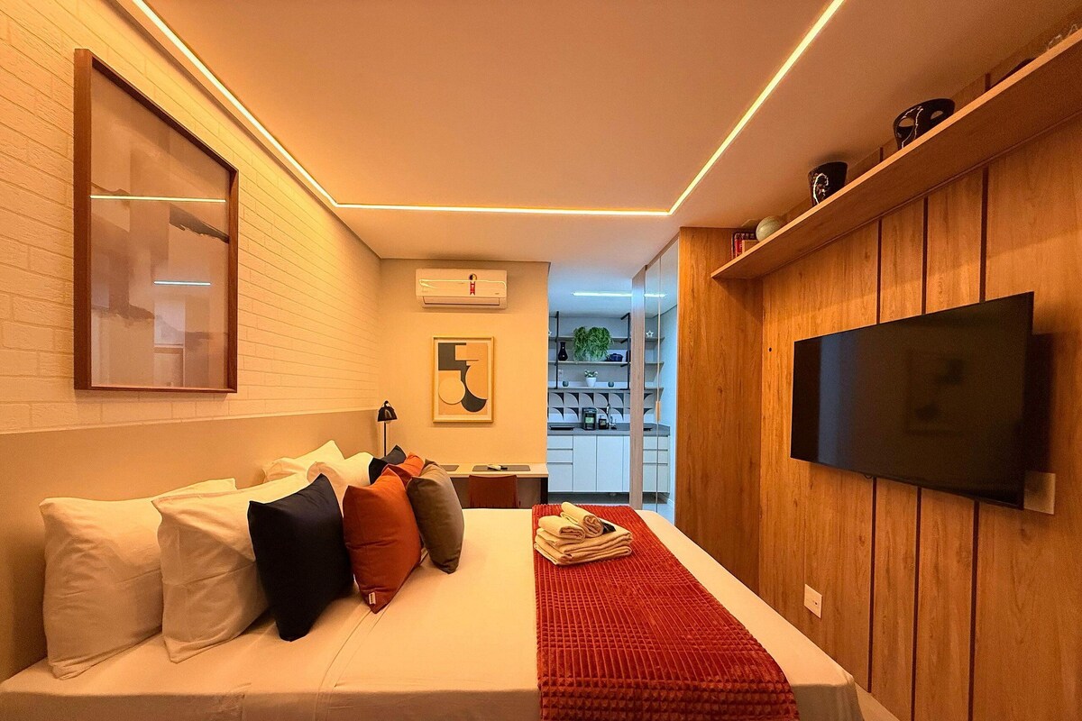 A modern hotel room with a bed, a TV, a small desk, and a kitchenette.