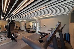 A gym with exercise equipment, a mirror, and a ceiling with linear light fixtures. - Condomínio Helbor Edition  (São Paulo)