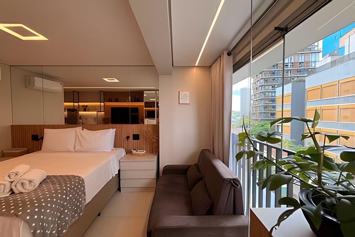 A modern hotel room with a bed, a sofa, a small table, a mirror, and a view of the cityscape.