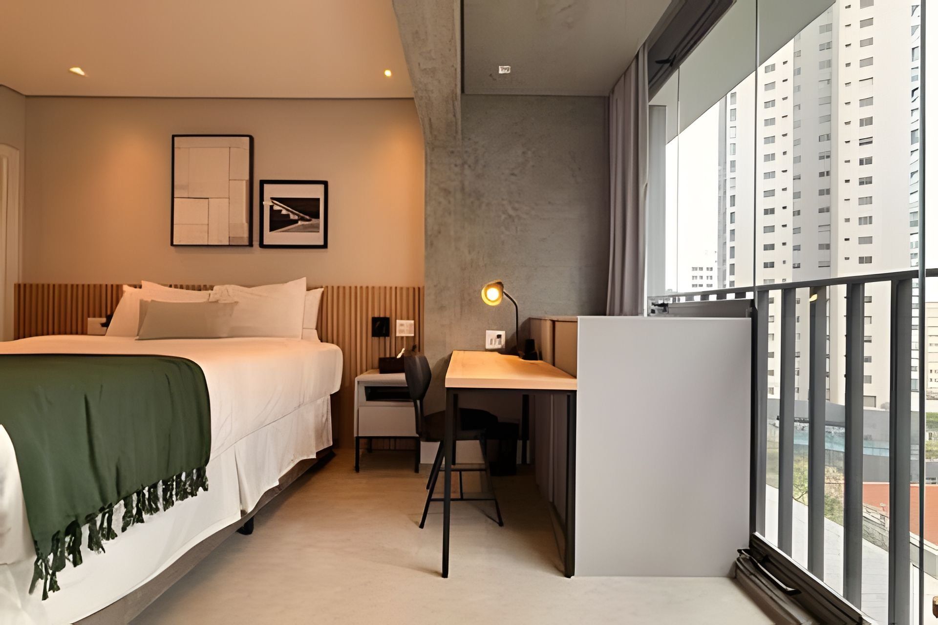 A hotel room with a bed, desk, chair, and large windows offering a view of high-rise buildings.