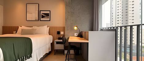 Habitación de hotel con cama, escritorio, silla y ventanas amplias que ofrecen vista a edificios altos.